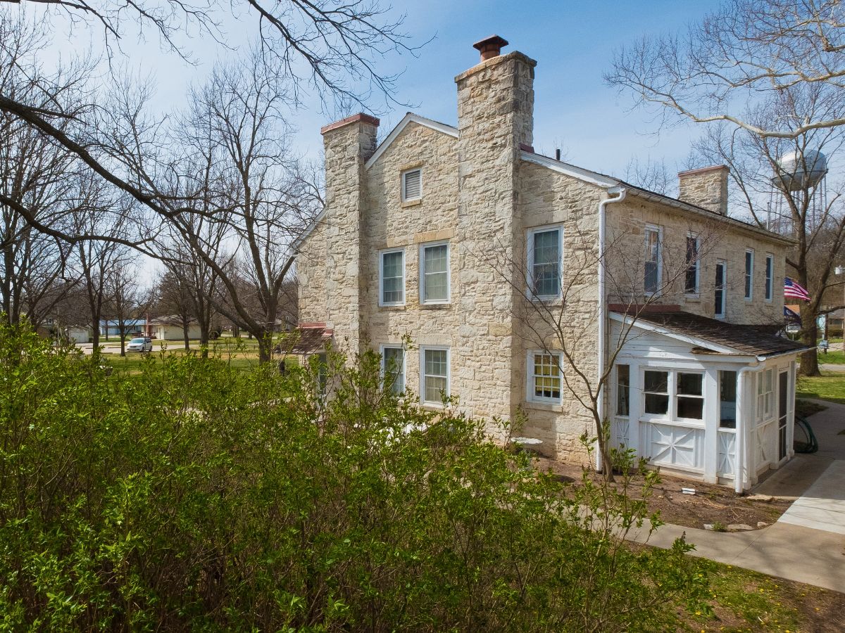 A historic stone house with chimneys, surrounded by trees and greenery, under a clear sky. An American flag is visible near the building.
