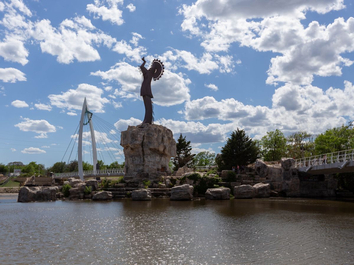 A tall statue stands on a rocky platform in the middle of a pond with a bridge and modern sculpture in the background, under a partly cloudy sky.