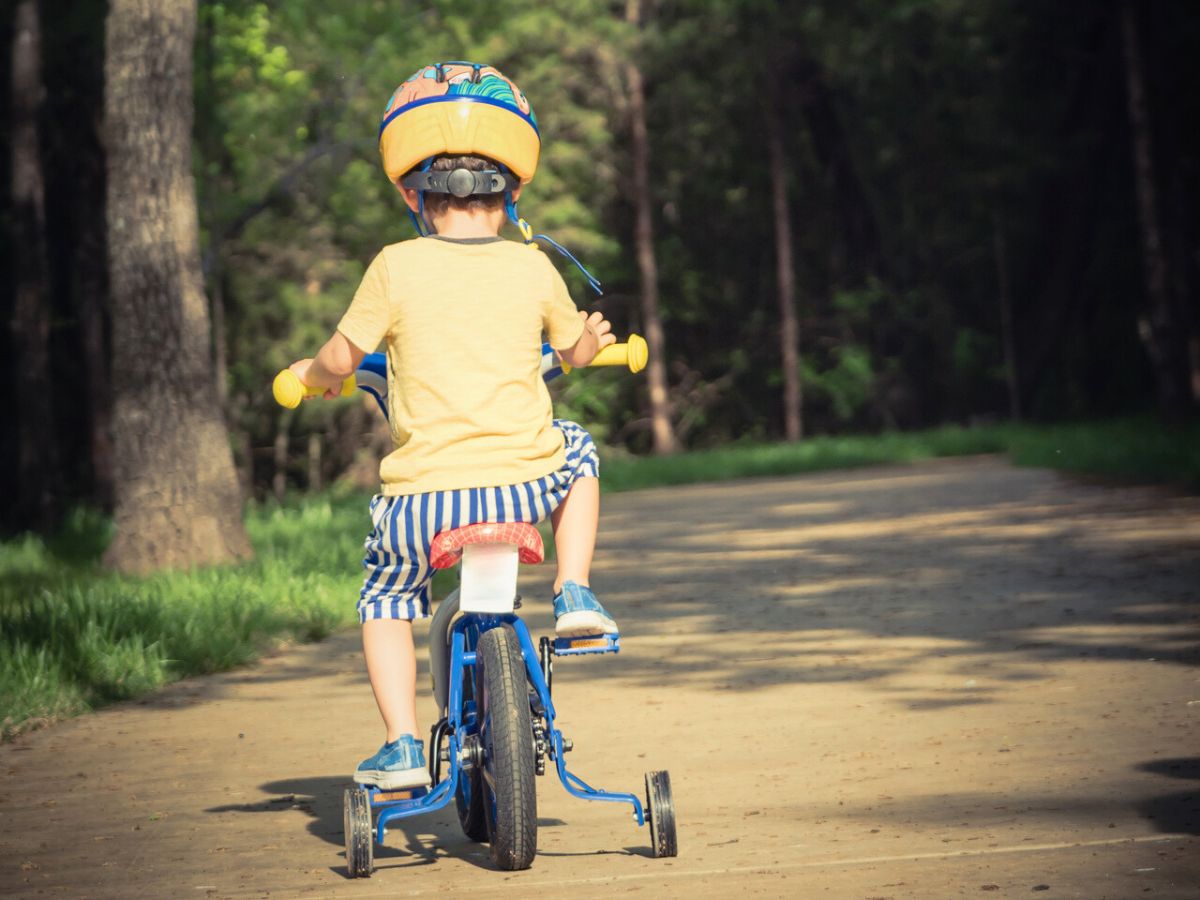 A child wearing a helmet and striped shorts is riding a blue tricycle on a forest path.