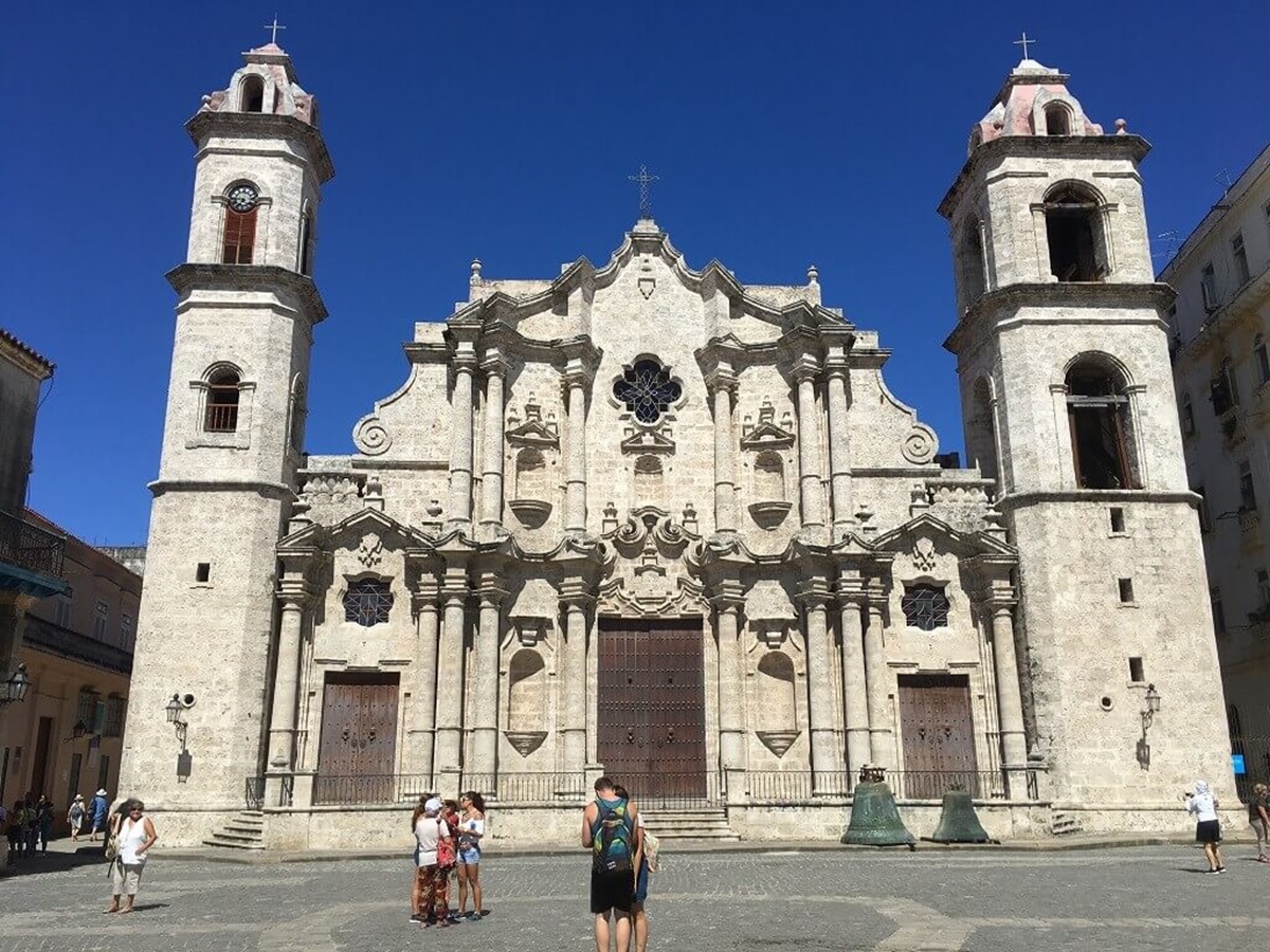 A group of tourists stands in front of a historic stone cathedral with twin bell towers and ornate details under a clear blue sky.