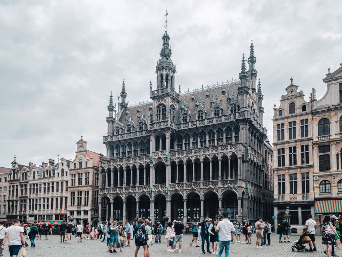 A crowd of people stands in front of a large, ornate building with Gothic architecture on a cloudy day, surrounded by similarly styled buildings.