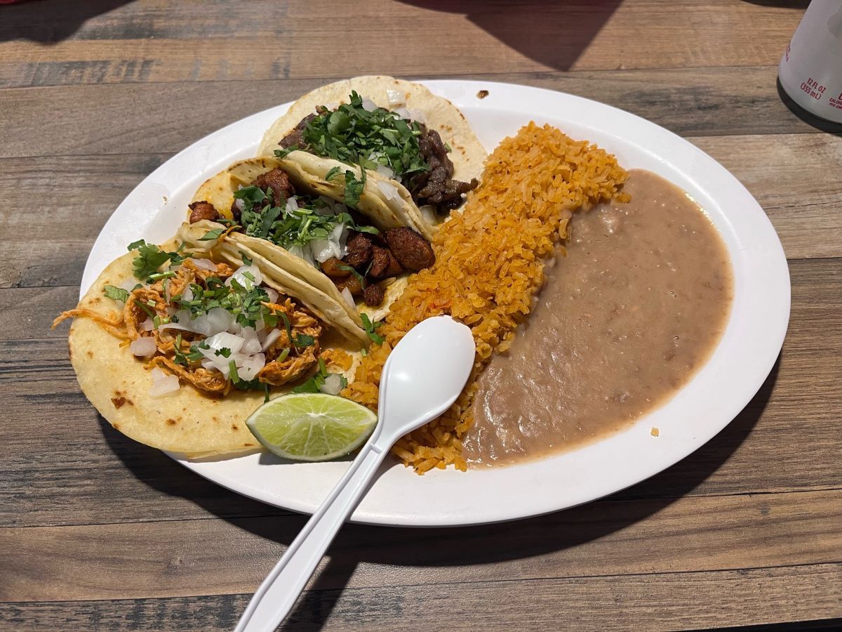 A plate of three tacos with cilantro and onions, served with a side of refried beans and Mexican rice. A plastic spoon and a wedge of lime are placed on the plate.