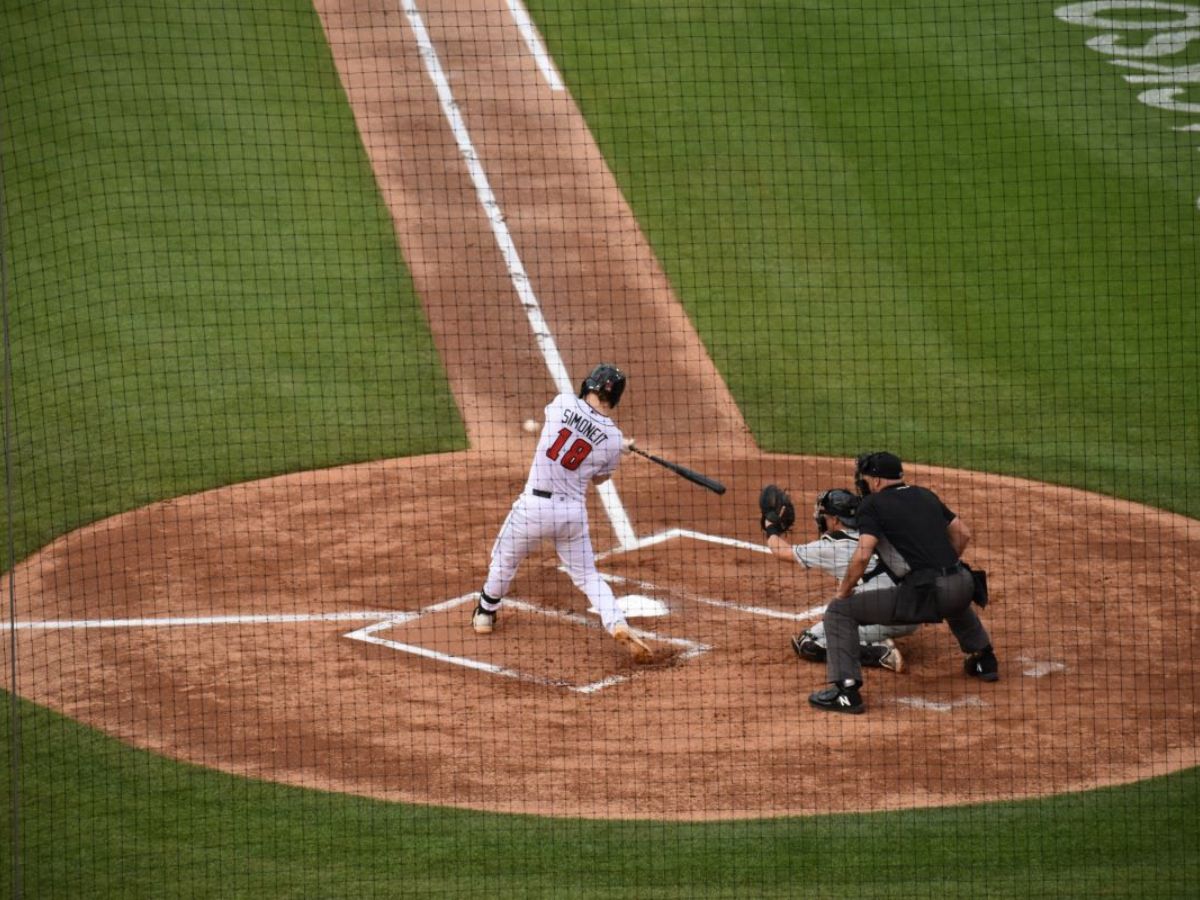 Baseball player in white uniform, number 18, swinging bat at home plate as catcher and umpire observe, during a game.
