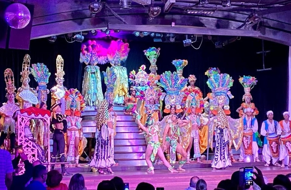 A vibrant stage performance featuring performers in elaborate, colorful costumes and headdresses. They dance on a multi-level platform, with a colorful backdrop and audience members watching.