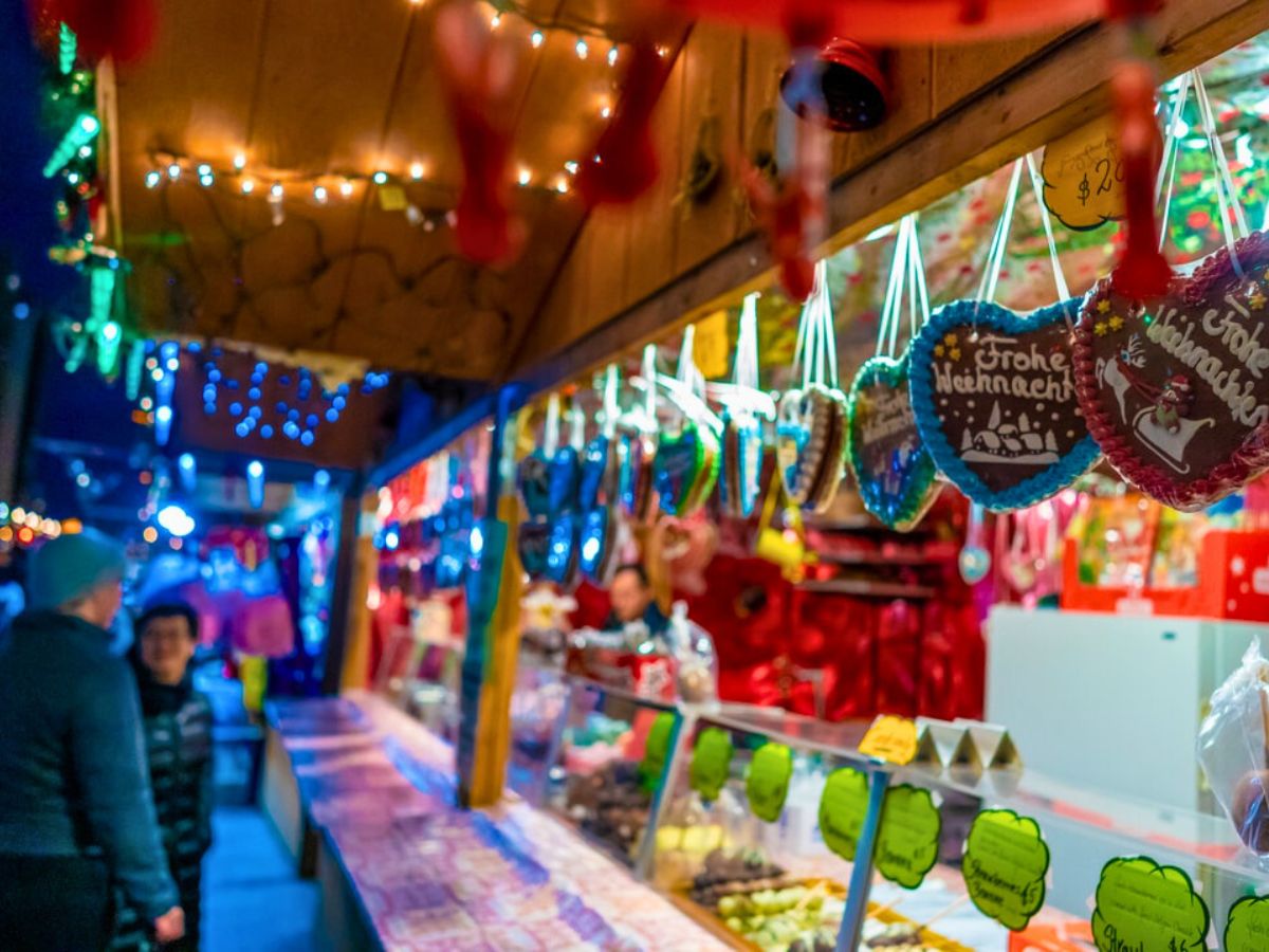 A festive market stall with colorful lights displays holiday-themed gingerbread cookies. Two people are visible in the foreground.