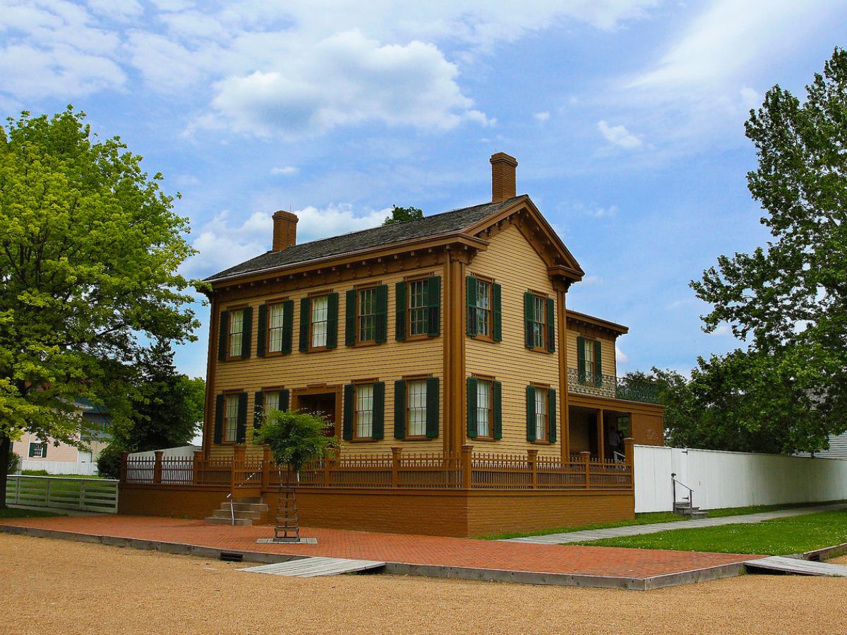 A two-story, brown wooden house with green shutters and a white fence in the background, set on a sunny day with a partly cloudy sky.