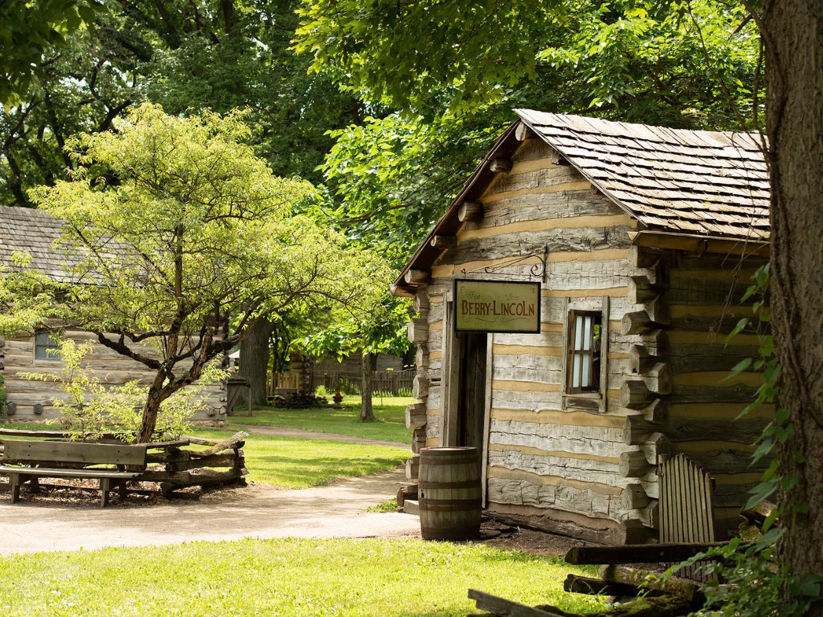 A small log cabin labeled "Berry-Lincoln" is nestled among trees and greenery on a bright sunny day. Another log building and a wooden bench are visible in the background.