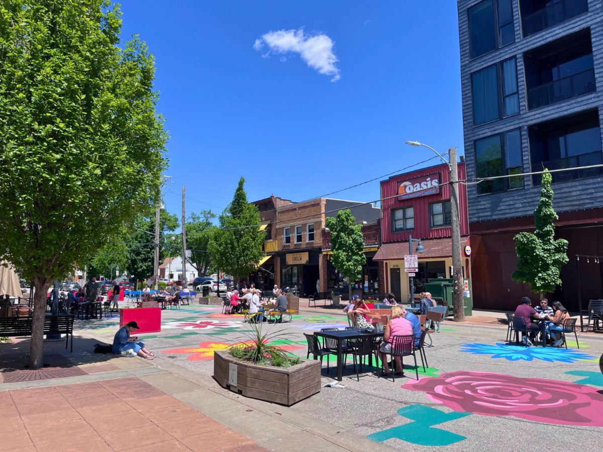 A vibrant street scene with colorful street art, outdoor seating, and people dining. Buildings and trees line the street under a clear blue sky.