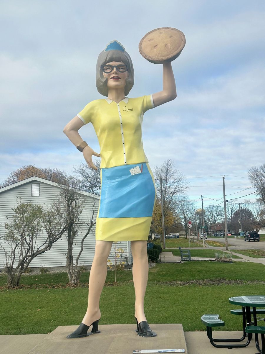 A large statue of a woman in a blue and yellow uniform holding a pie aloft, standing on a concrete base. The background features a grassy area, trees, and a few houses.