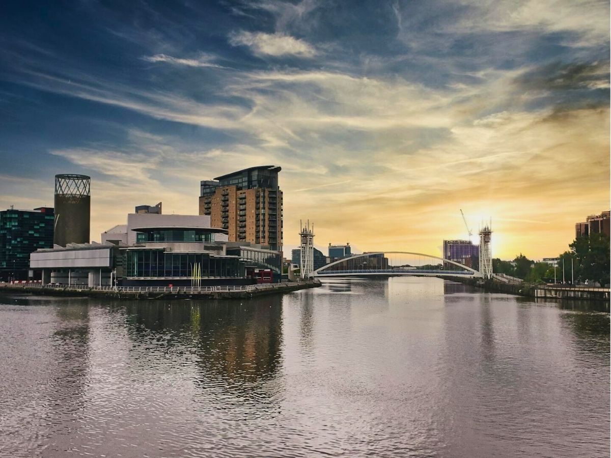 A river flows through a cityscape with various buildings and a bridge under a sky transitioning from day to sunset.