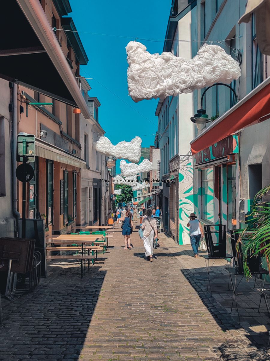 Cobblestone street lined with shops and outdoor seating, with several fluffy cloud-like art installations hanging above. People are walking and some are sitting under clear blue skies.