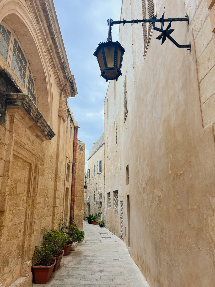 A narrow, stone-paved alley with potted plants lining the base of the walls and an old-fashioned lantern hanging from a wrought iron bracket on the right. The walls are light-colored and weathered.