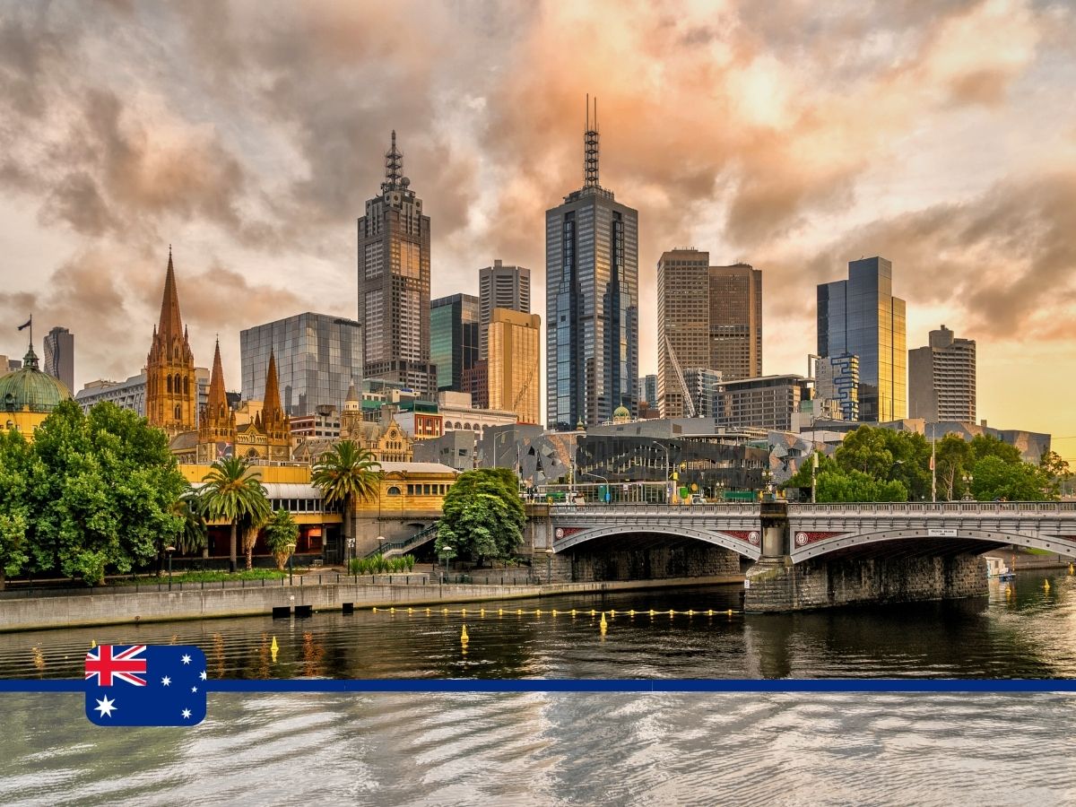Skyline of Melbourne, Australia, featuring modern skyscrapers and historic buildings at sunset, with the Yarra River in the foreground and an Australian flag at the bottom left corner.