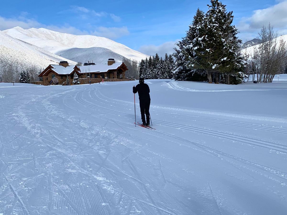 A person cross-country skiing on a snowy landscape with a lodge and trees in the background.