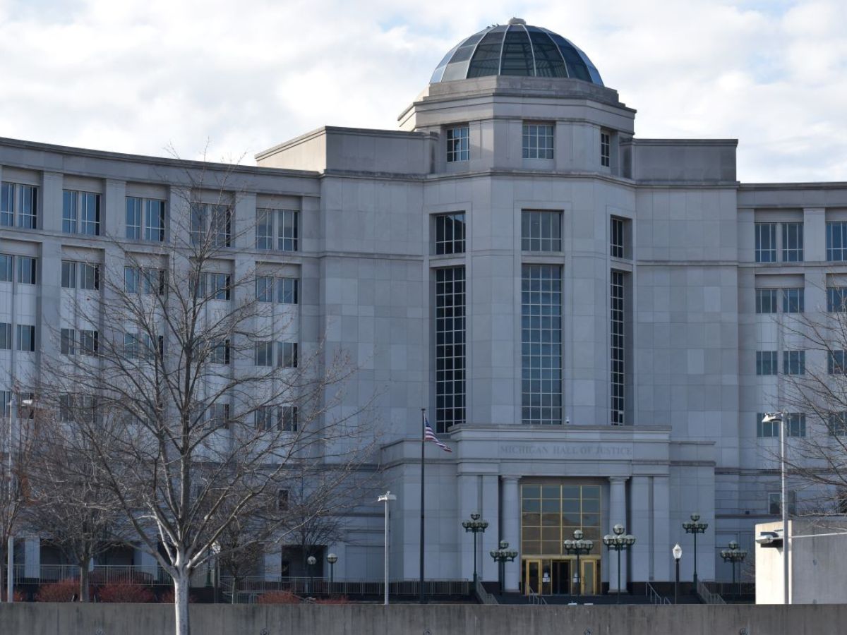 The image displays the front facade of the Michigan Hall of Justice, a large gray building with a central dome, multiple windows, and an American flag near the entrance.