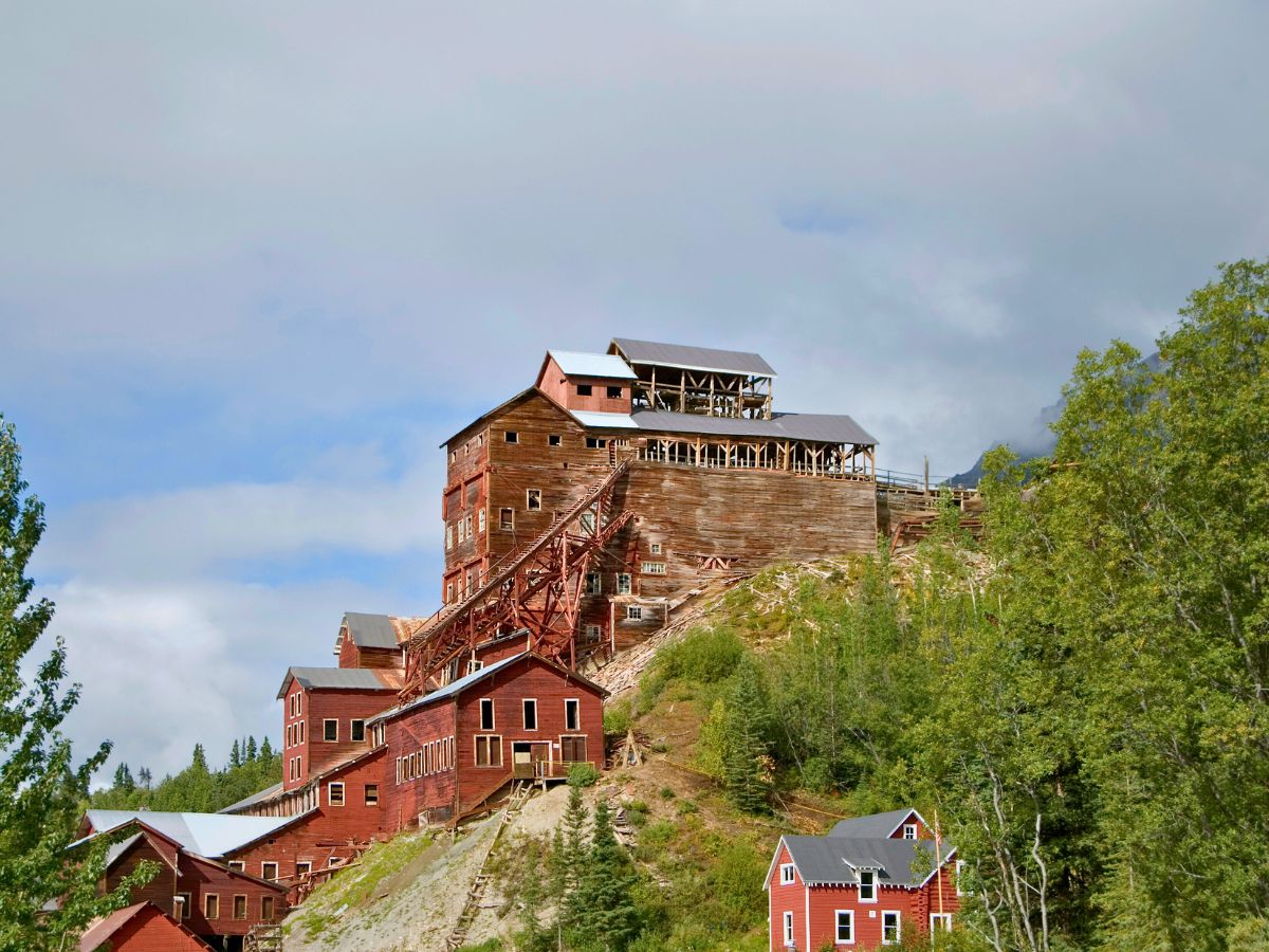 Historic wooden mining building with multiple levels and staircases sits on a hill surrounded by greenery and cloudy sky.