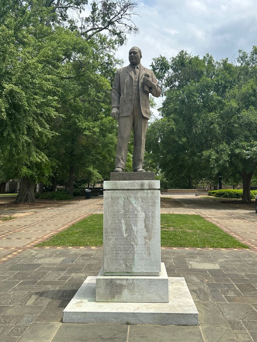 A statue of Martin Luther King Jr. stands on a stone pedestal, surrounded by trees and a paved walkway in a park.