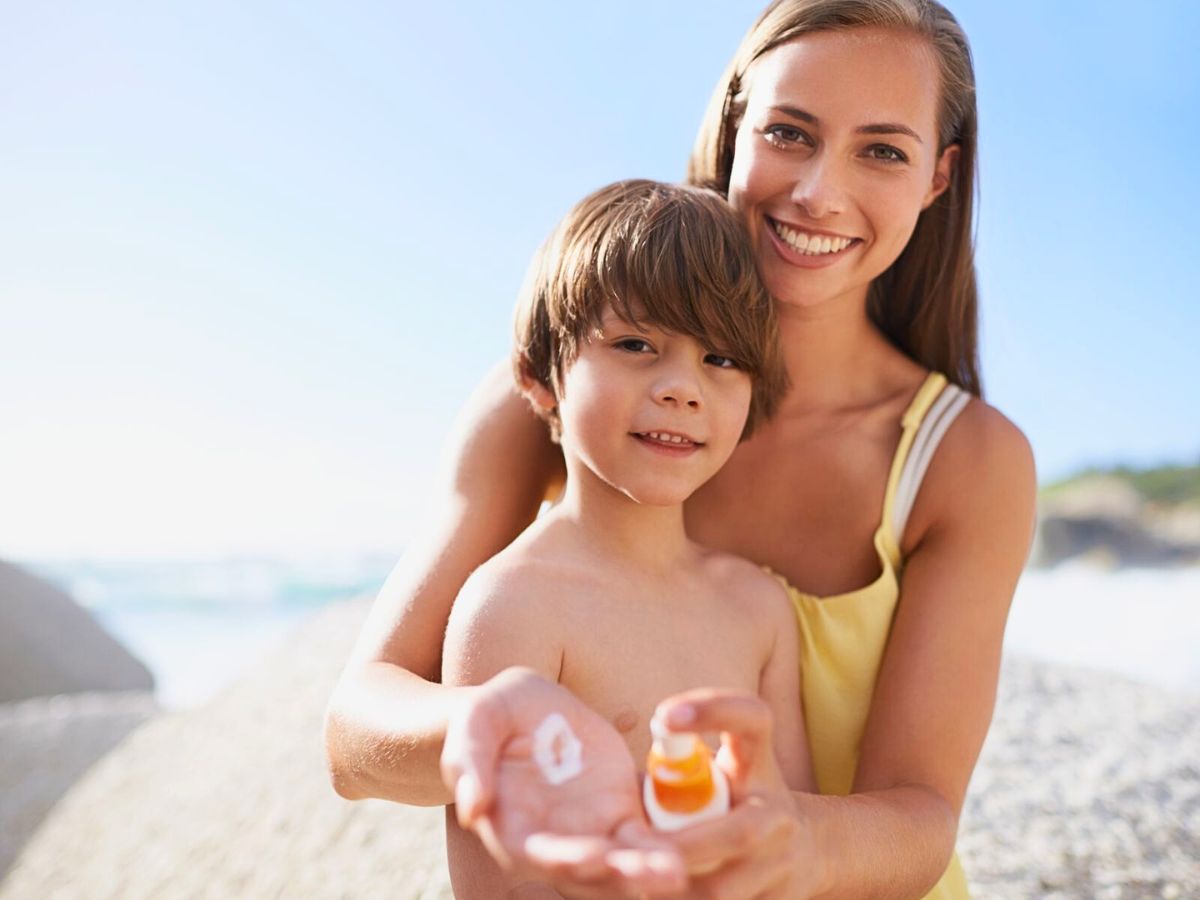 A woman and a child with a sunscreen bottle on the beach, both smiling at the camera. The woman is holding some sunscreen in her hand, ready to apply it.
