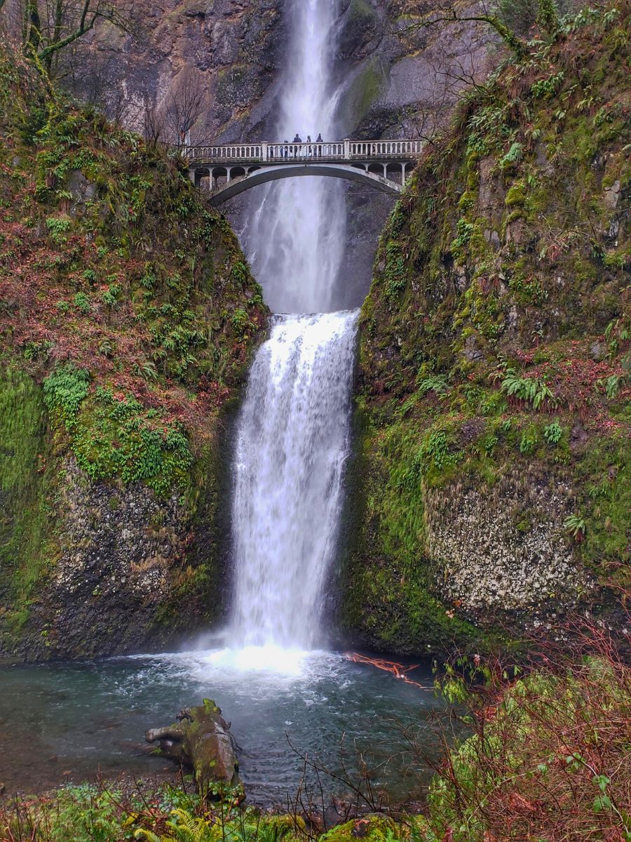 A two-tiered waterfall cascades beneath a stone bridge surrounded by lush green vegetation.