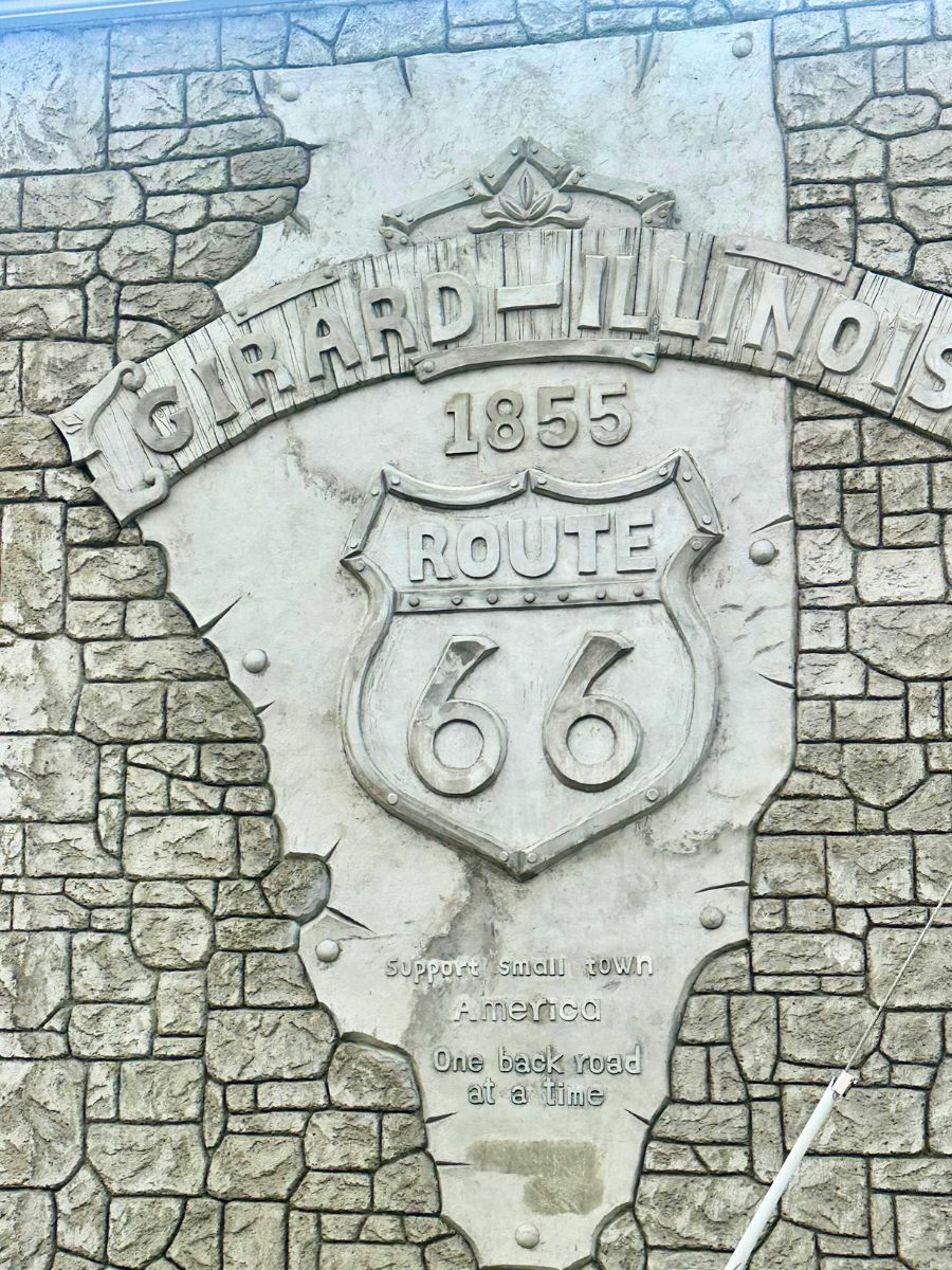 Stone mural of Girard, Illinois, featuring Route 66 emblem and text: "Support small town America, one back road at a time." Engraved alongside a stone brick pattern.