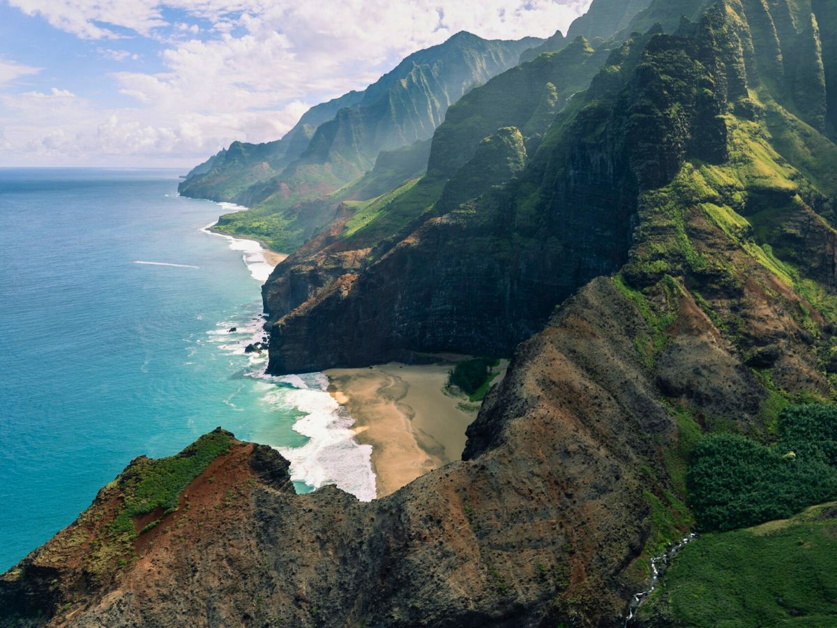 Aerial view of a rugged coastline with steep cliffs, green vegetation, and turquoise ocean under a partly cloudy sky.