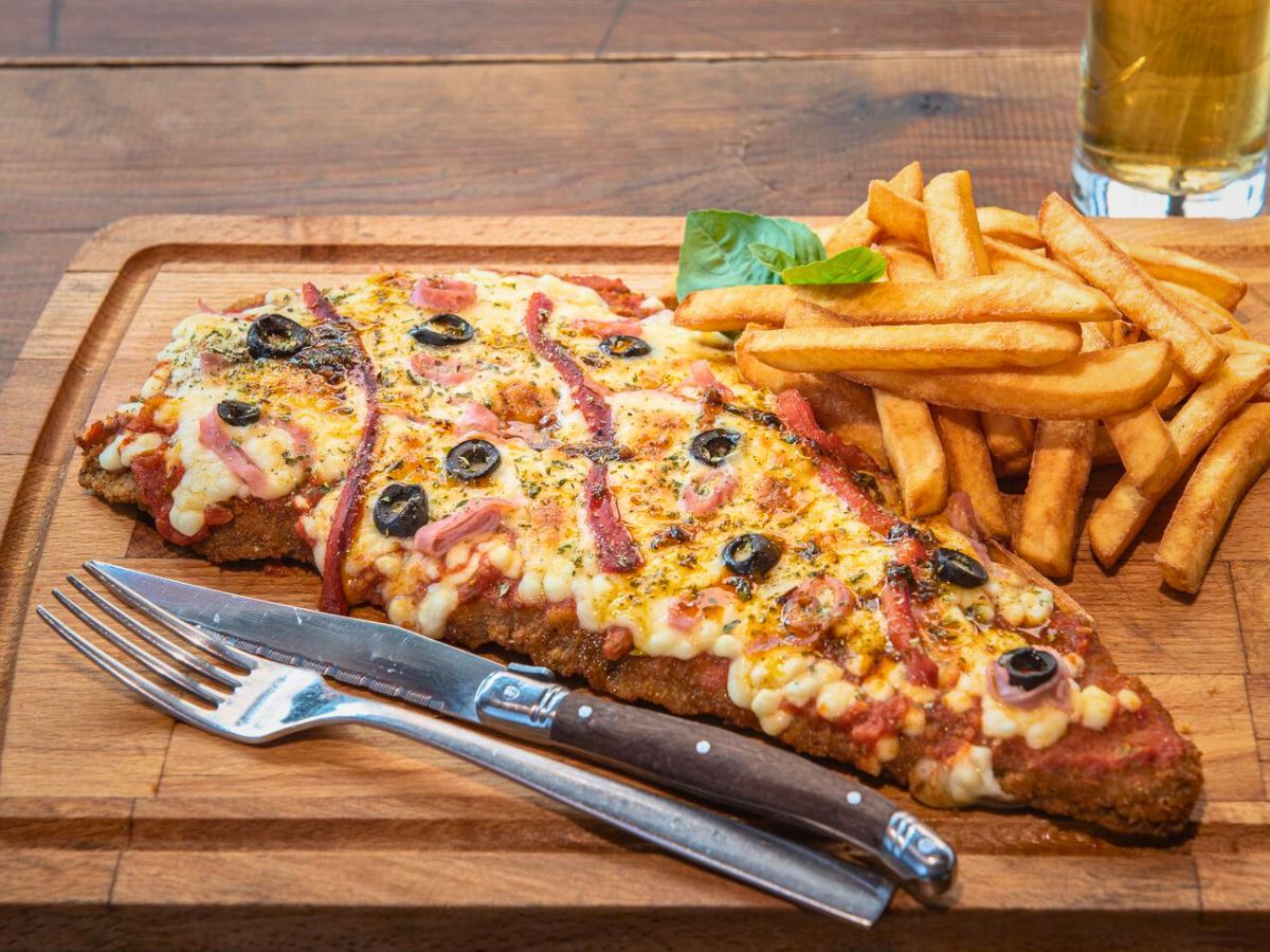 A breaded and fried piece of meat topped with melted cheese, olives, and tomato sauce, served with a side of French fries on a wooden board, with a knife and fork placed beside them.