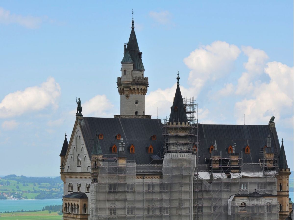 Neuschwanstein Castle with scaffolding on part of the building, set against a backdrop of a cloudy sky and green landscape.