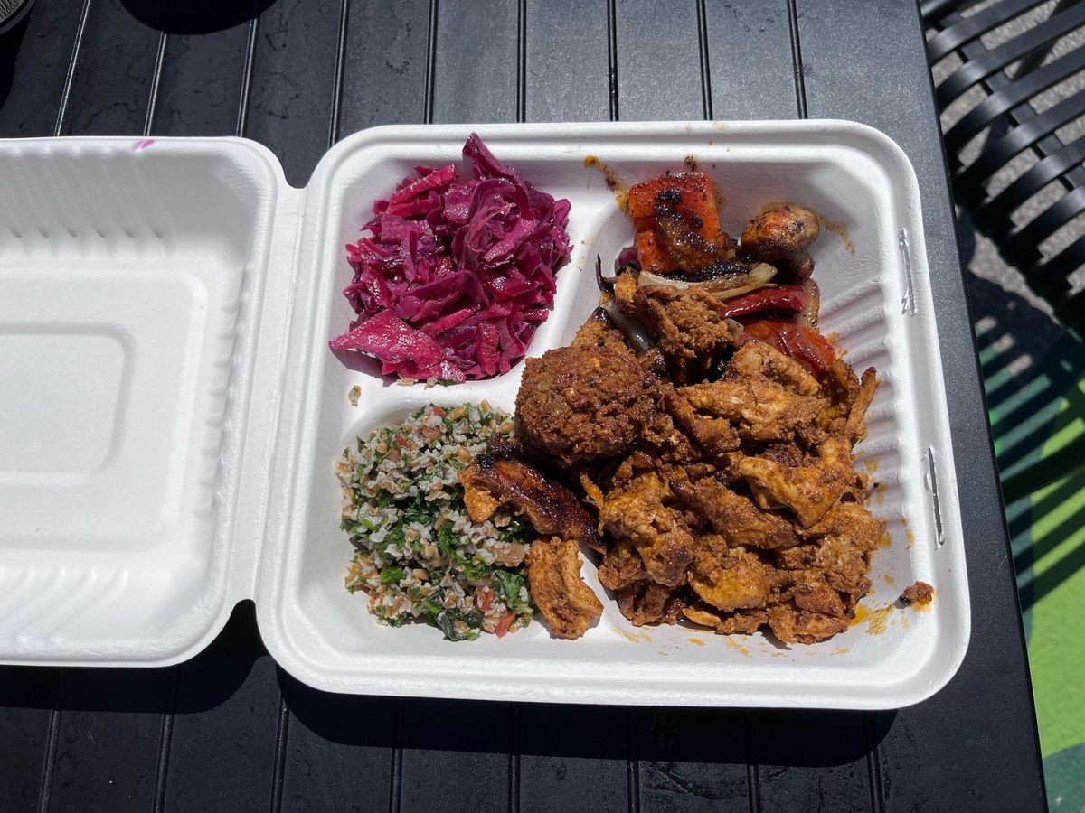 A takeout container with falafel, spiced chicken, grilled vegetables, tabbouleh, and red cabbage slaw, placed on an outdoor table.