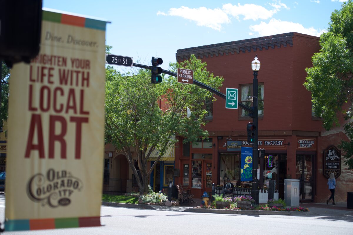 Street corner with a red brick building, trees, and shops. A "Discover Local Art" banner and a "Public Parking" sign are visible. Clear sky in the background.