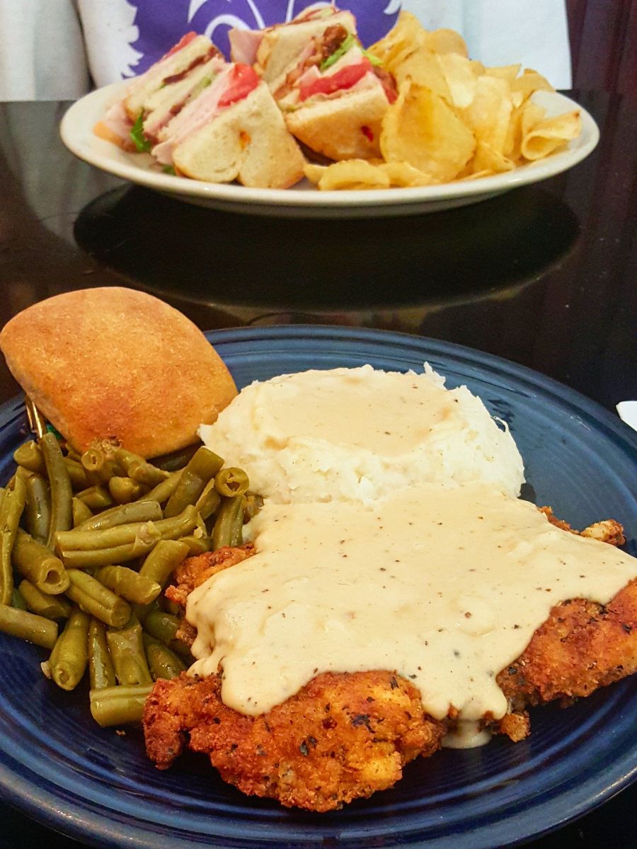 A blue plate with chicken fried steak topped with gravy, mashed potatoes, green beans, and a roll. In the background, there is a plate with a sandwich and potato chips.