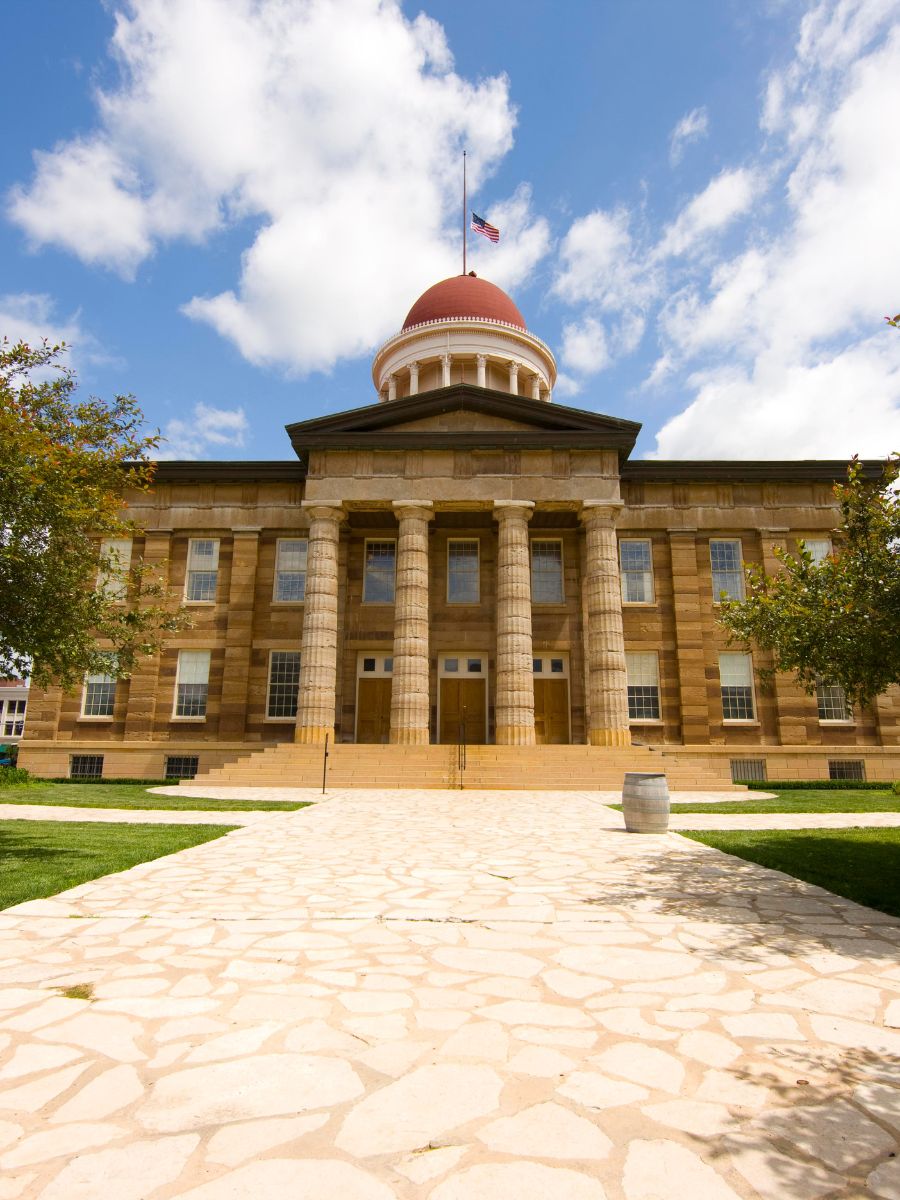 Historic courthouse building with a central domed tower, bordered by large stone columns, set against a sky with scattered clouds and surrounded by trees. Stone pathway leads to the entrance.