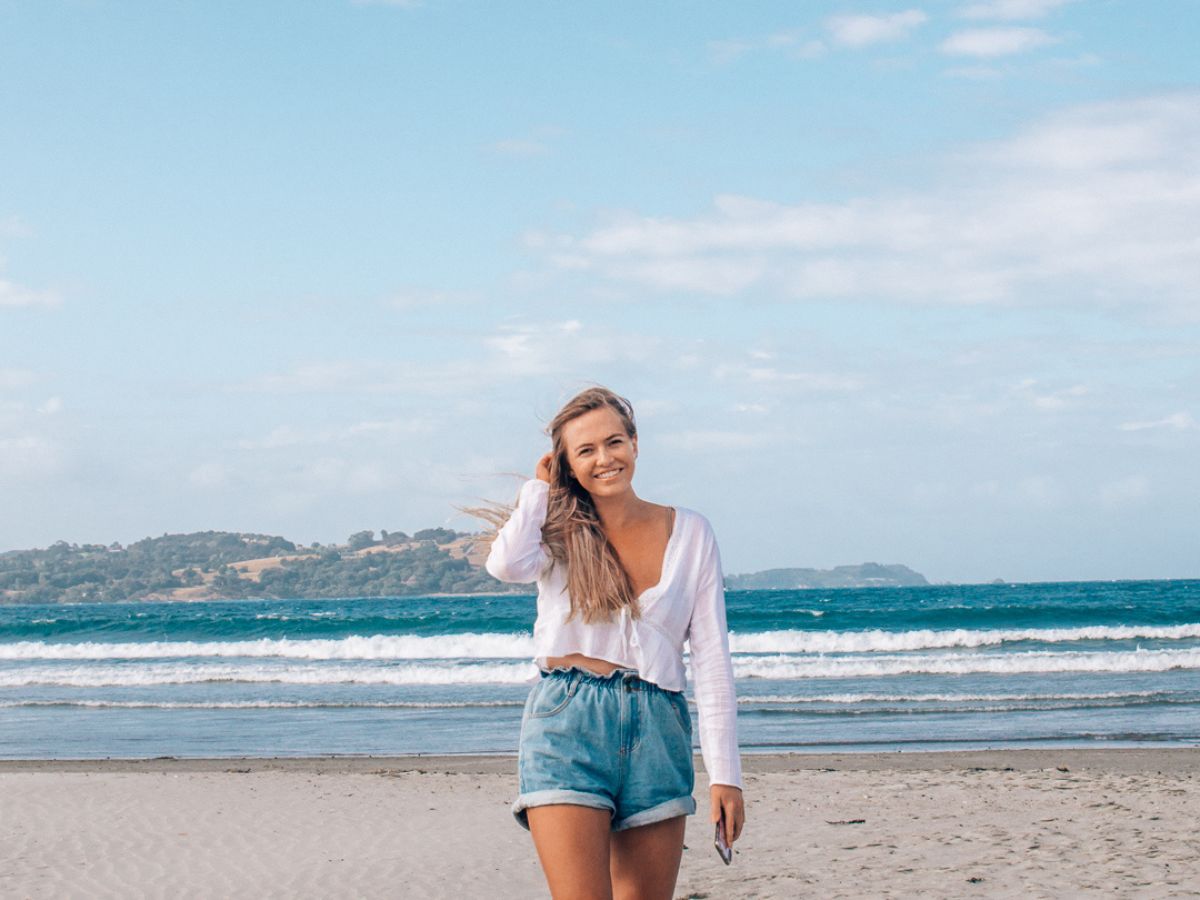 A woman wearing a white top and denim shorts stands on a beach, smiling, with ocean waves in the background and distant hills under a partly cloudy sky.