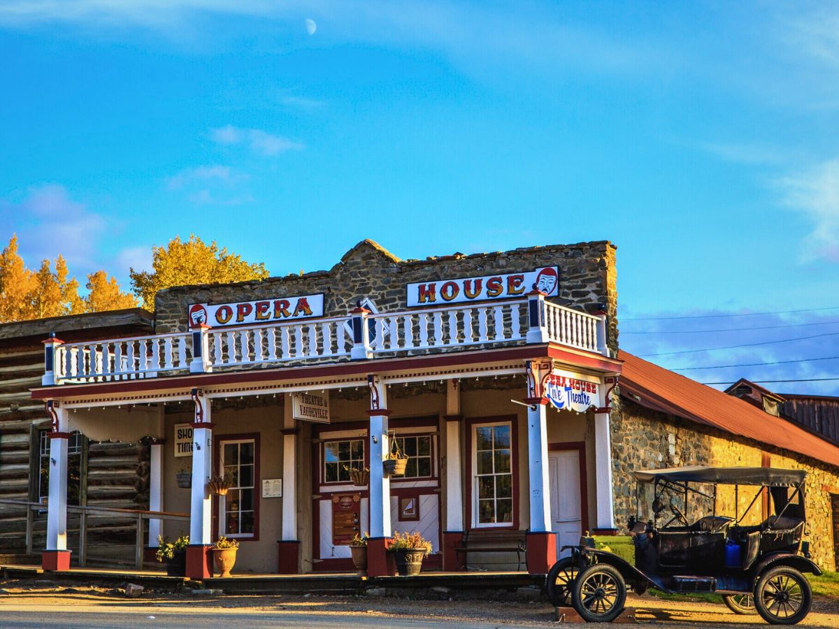 A vintage car is parked in front of a rustic opera house with a stone facade and white wooden balcony under a clear blue sky.
