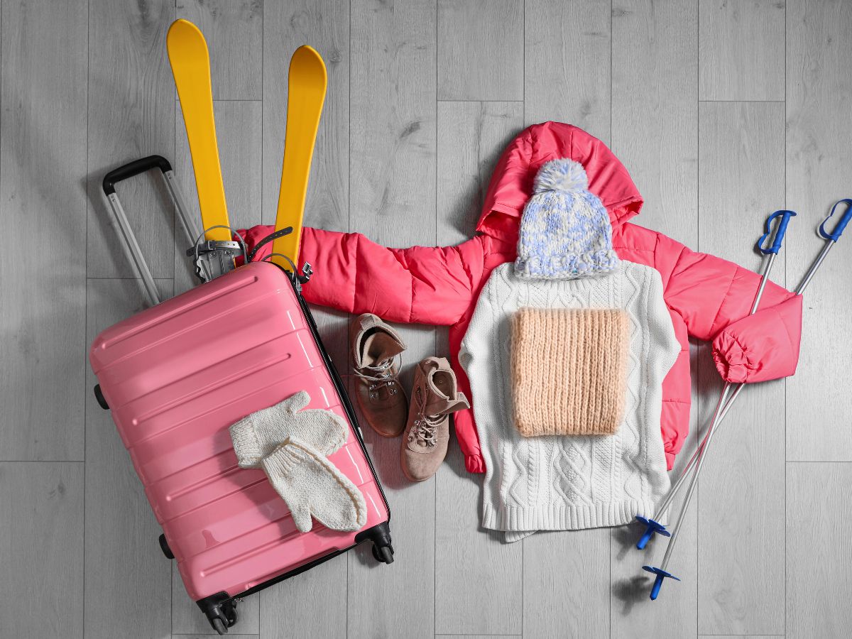 Pink suitcase, yellow skis, red jacket, white sweater, hat, gloves, brown boots, and ski poles arranged on a wooden floor.