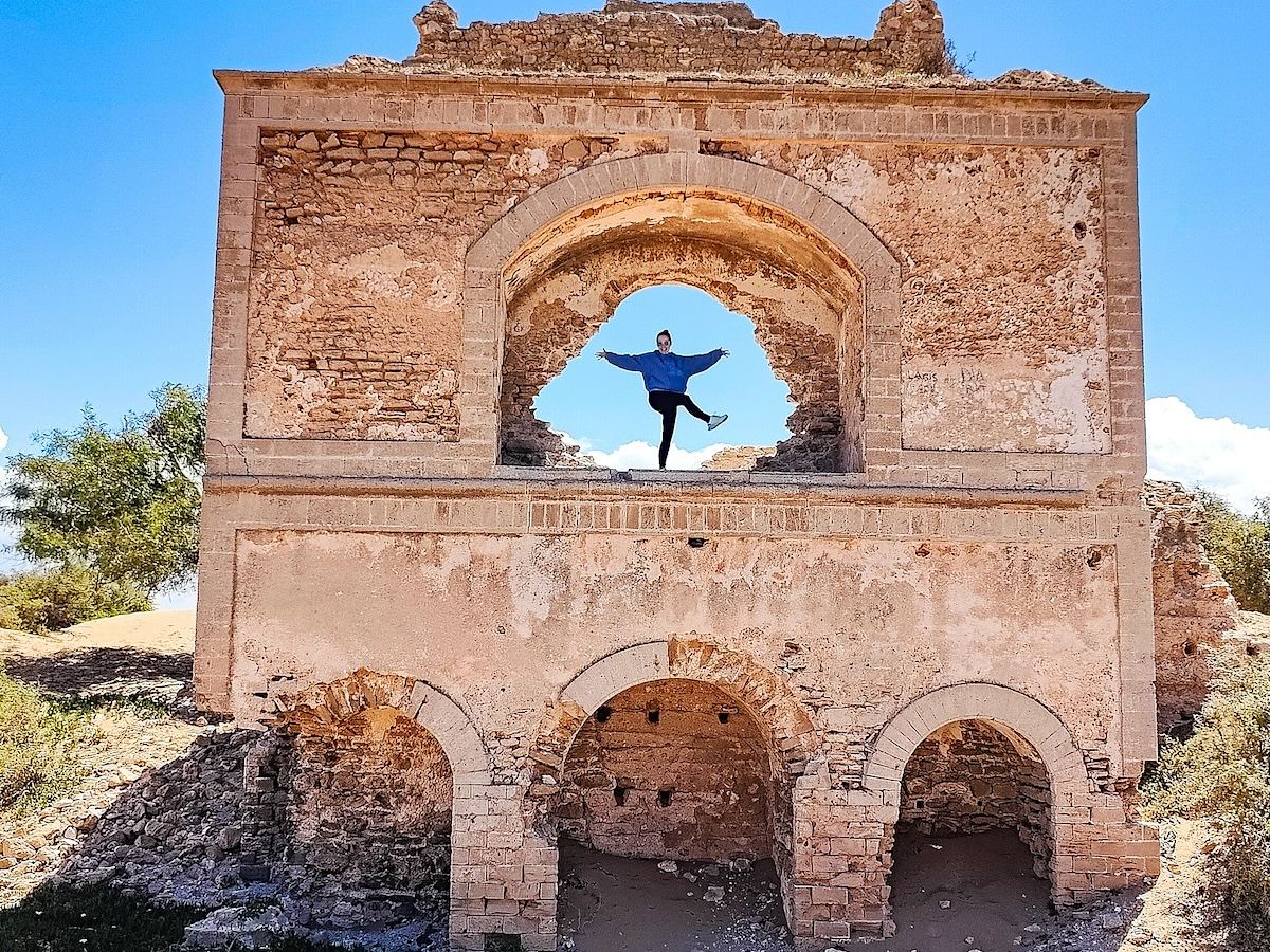 A person stands and stretches arms and legs in a framed archway of a dilapidated, historic brick structure under a blue sky.