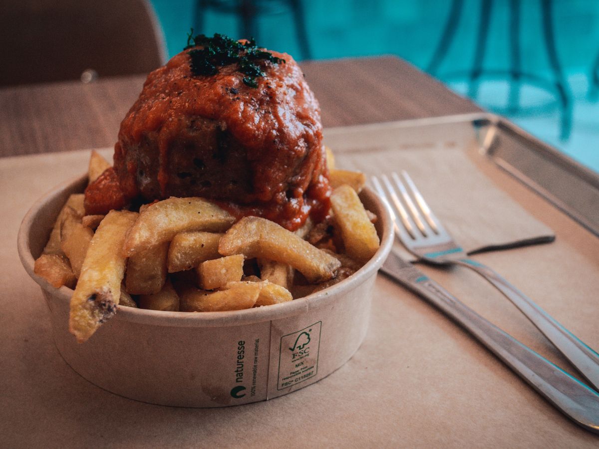 A bowl of fries topped with a meatball and red sauce is placed on a tray with a knife and fork beside it.