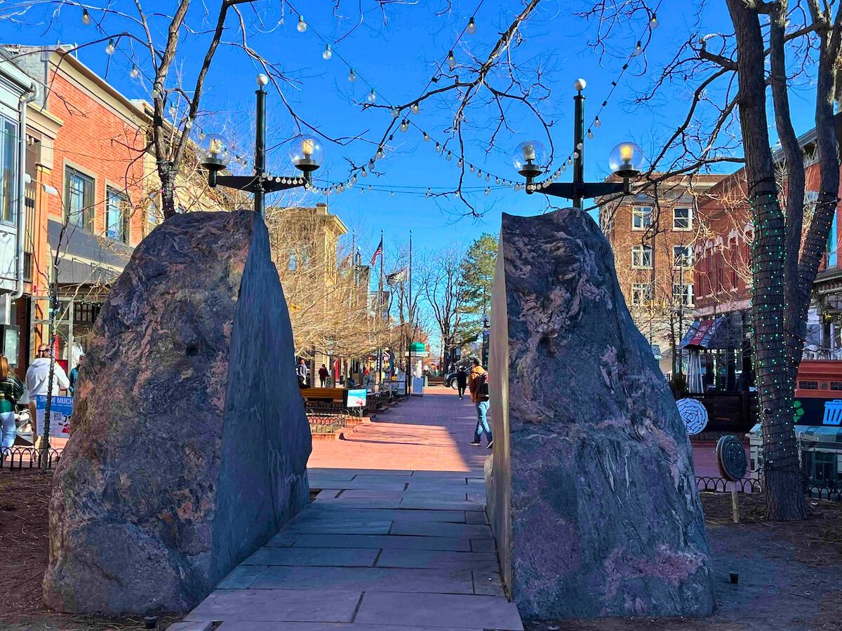 A pathway with large stone slabs on either side, surrounded by buildings and trees adorned with lights. People are walking in the distance.
