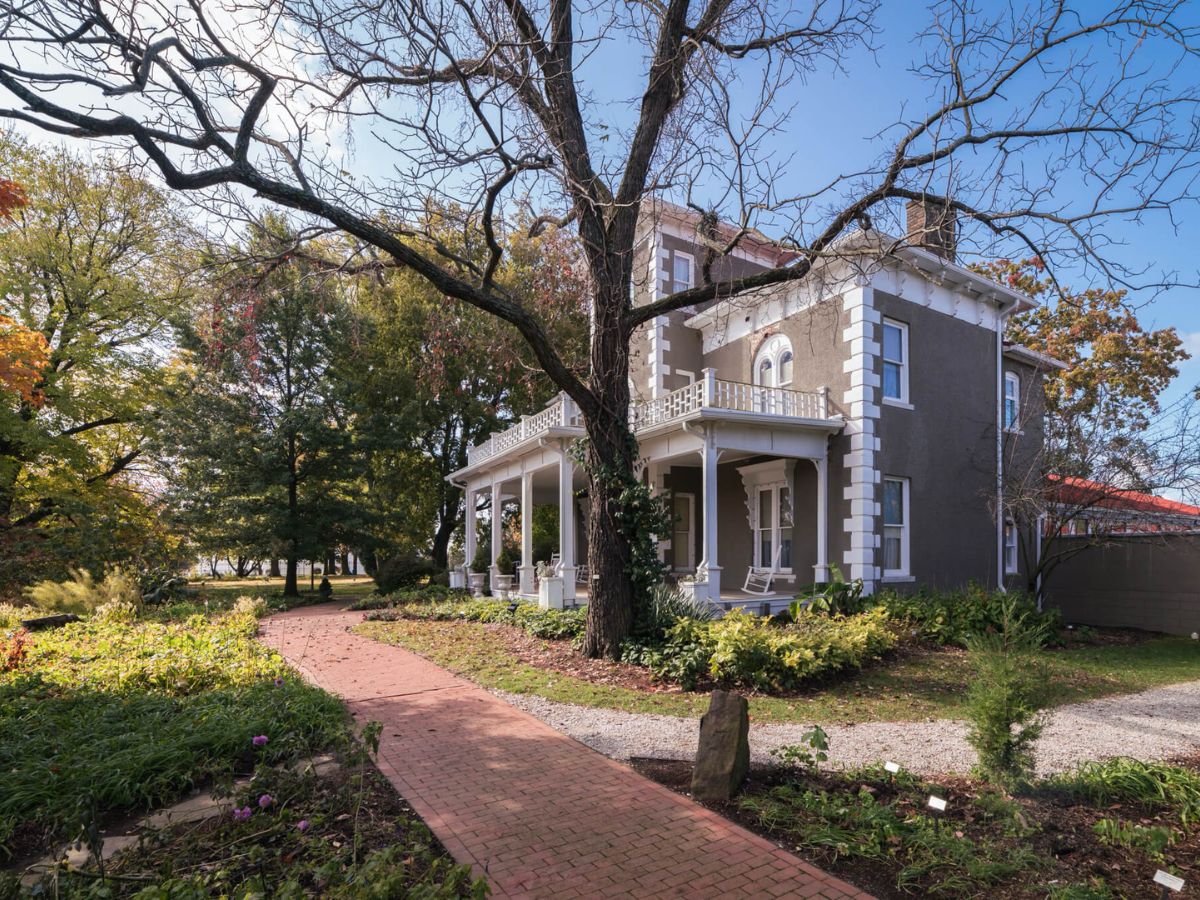 Two-story house with a large porch, decorative columns, and a brick pathway leading to the entrance, surrounded by trees and greenery on a sunny day.