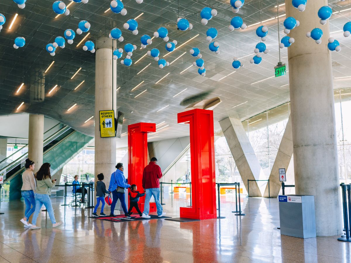 A family of five walks into a modern building with large red doorframes and blue spherical decorations hanging from the ceiling.