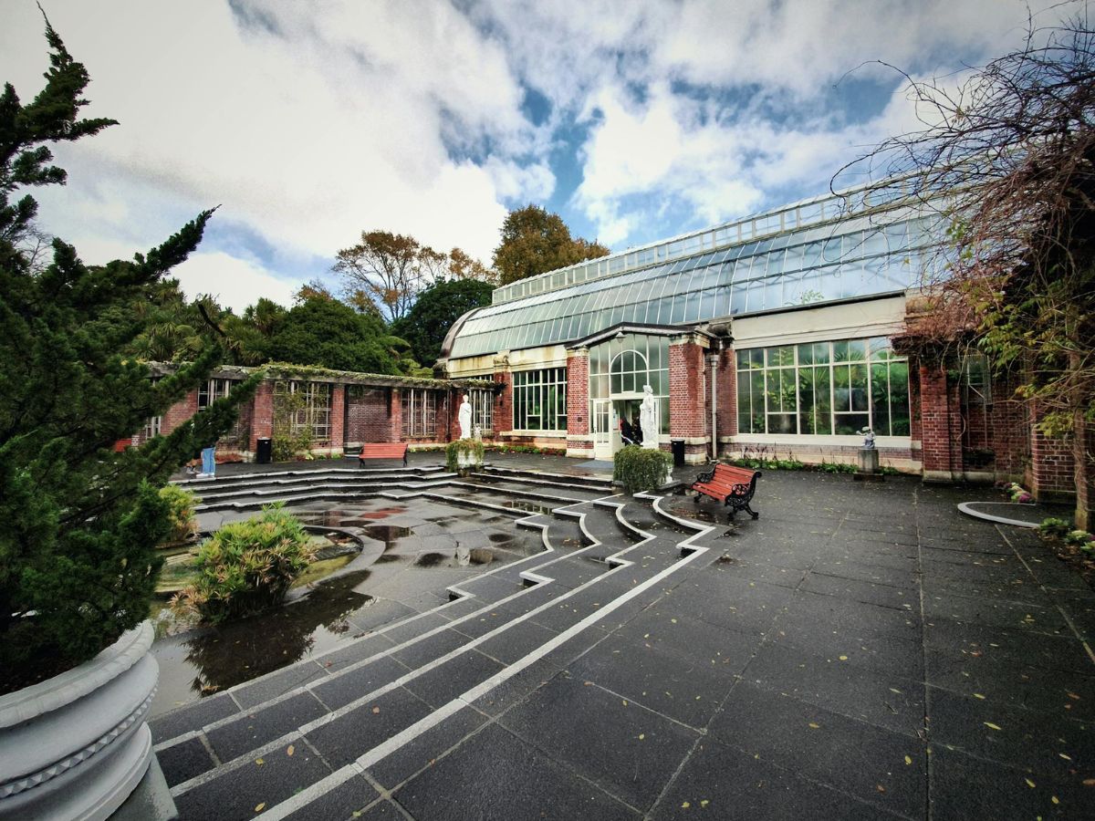 A large greenhouse with glass panels and brick walls surrounded by trees, bushes, and water features on a cloudy day, with a few benches and a paved walkway in the foreground.
