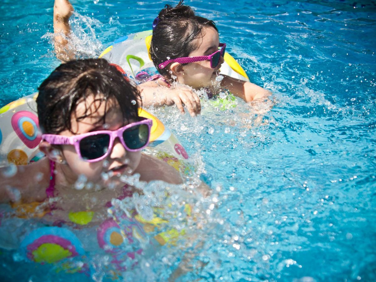 Two children wearing sunglasses and colorful swimsuits play in a pool, splashing water and using inflatable pool rings.