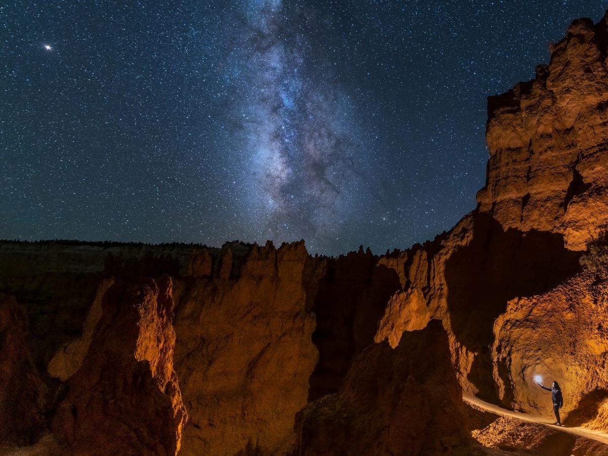 A person stands in a cave opening under a starry sky with the Milky Way visible above rocky formations.