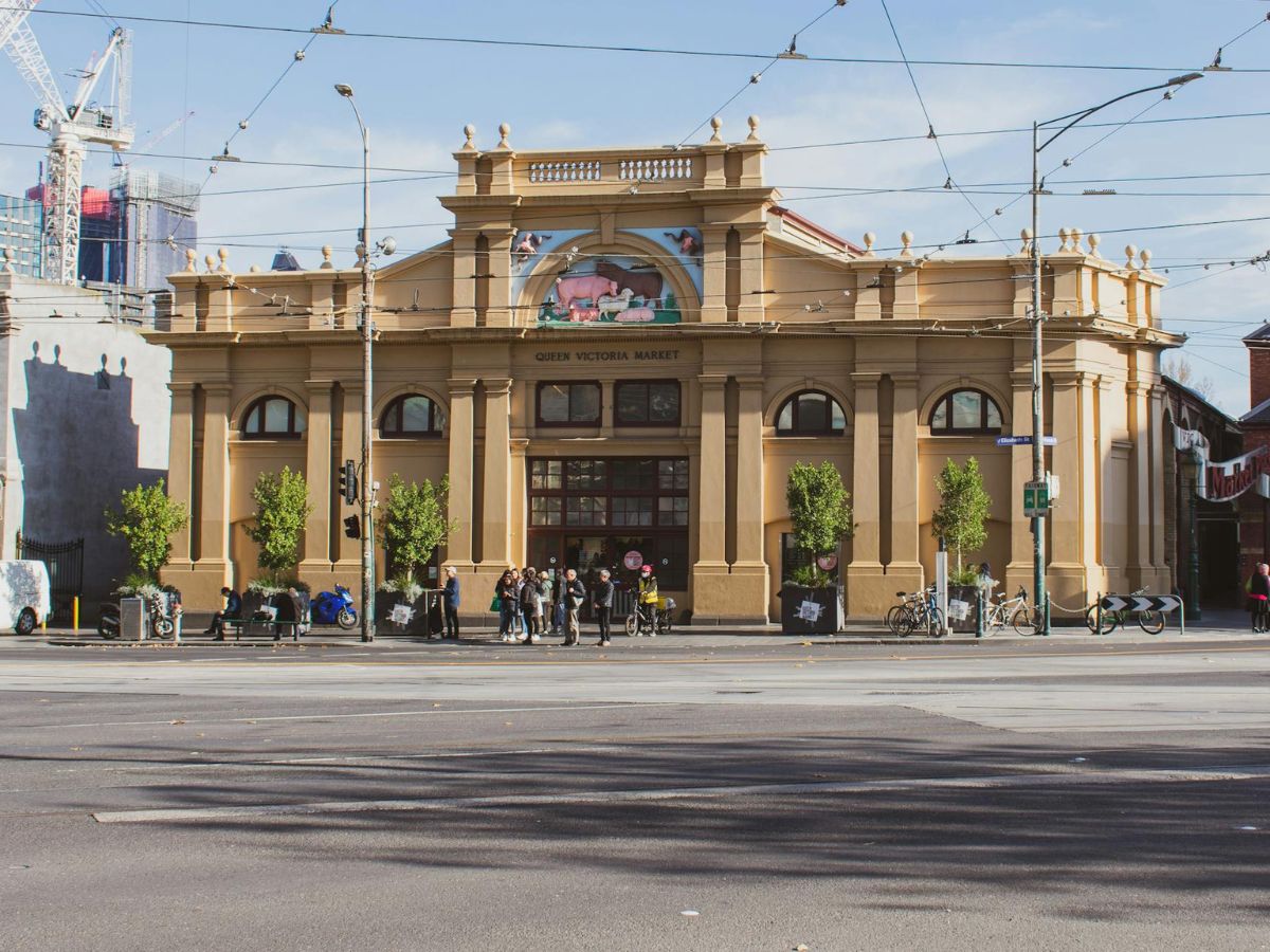 A wide street view of Queen Victoria Market in Melbourne, showing the historic building facade, overhead tram lines, and a small group of people gathered at the entrance.