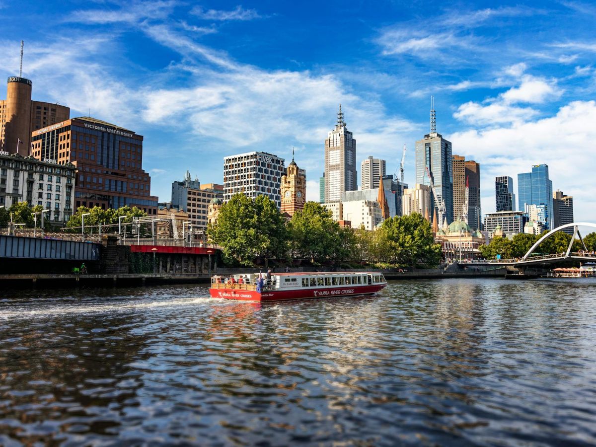 A red tour boat sails on a river with a backdrop of city buildings and a blue sky with some clouds. Trees line the riverbank.