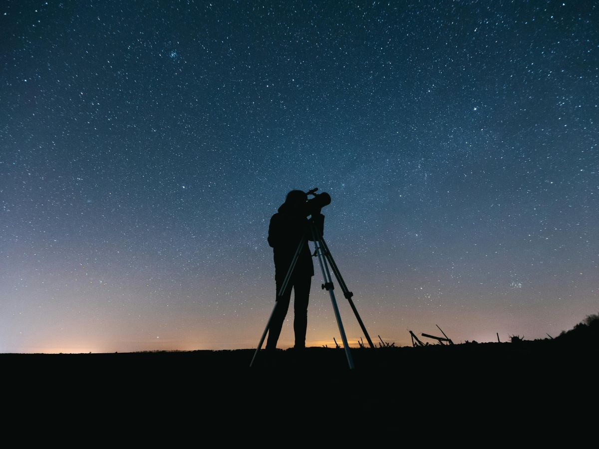 A person standing outdoors at night is looking through a telescope under a clear, starry sky. Silhouette and dark landscape contrast with the illuminated night sky.
