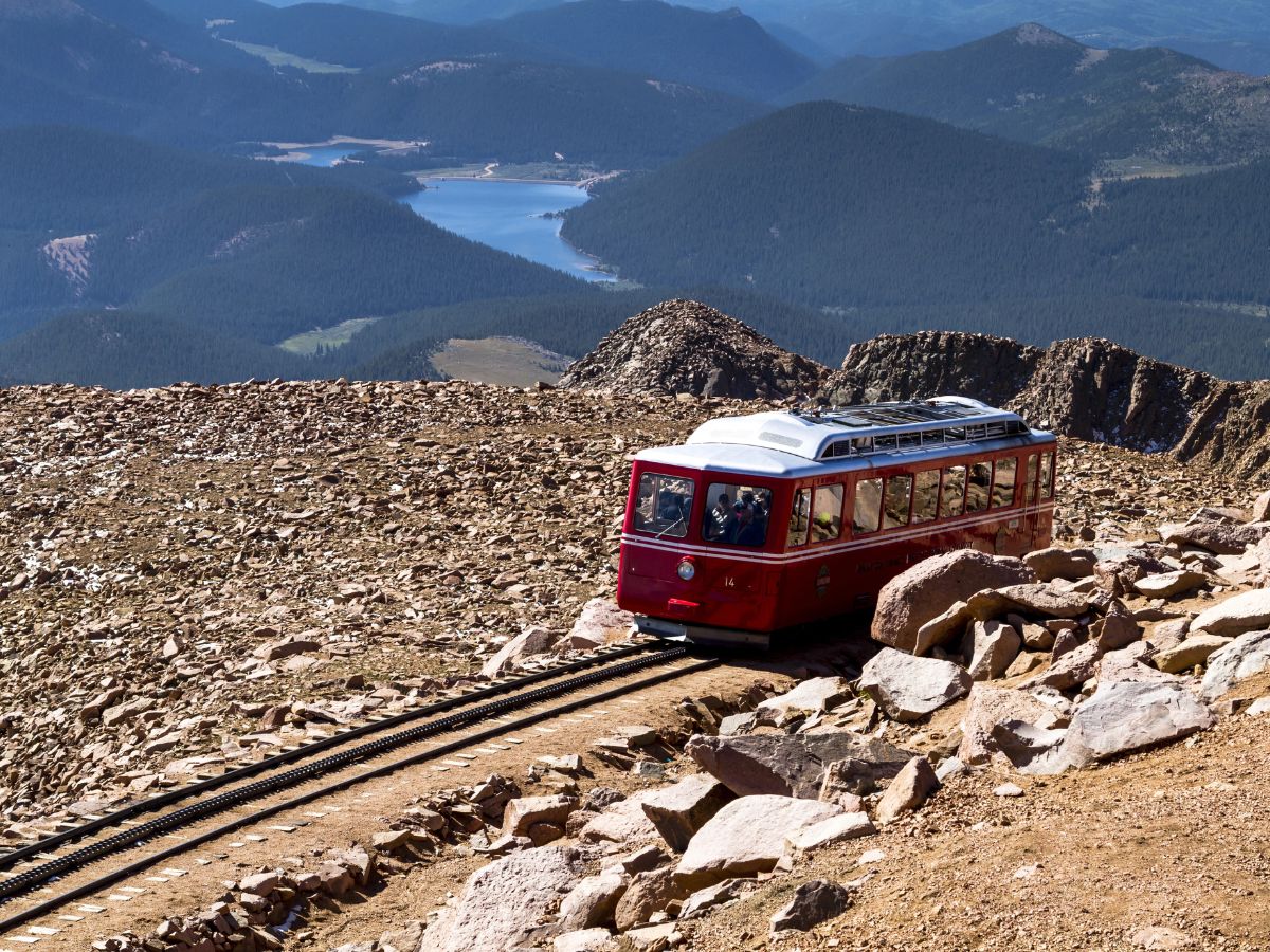 A red tram ascends a rocky mountain, with distant views of a lake and forested hills under a clear sky.
