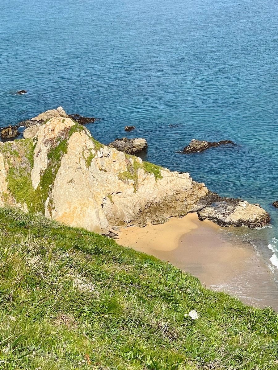 A rocky cliff with a grassy top overlooks a small sandy beach with clear blue water and scattered rocks.
