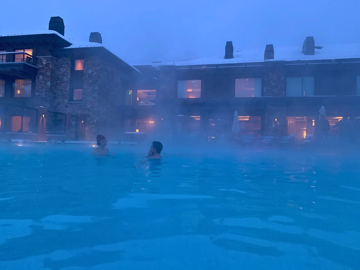 Two people swimming in a misty outdoor pool near a building at dusk.