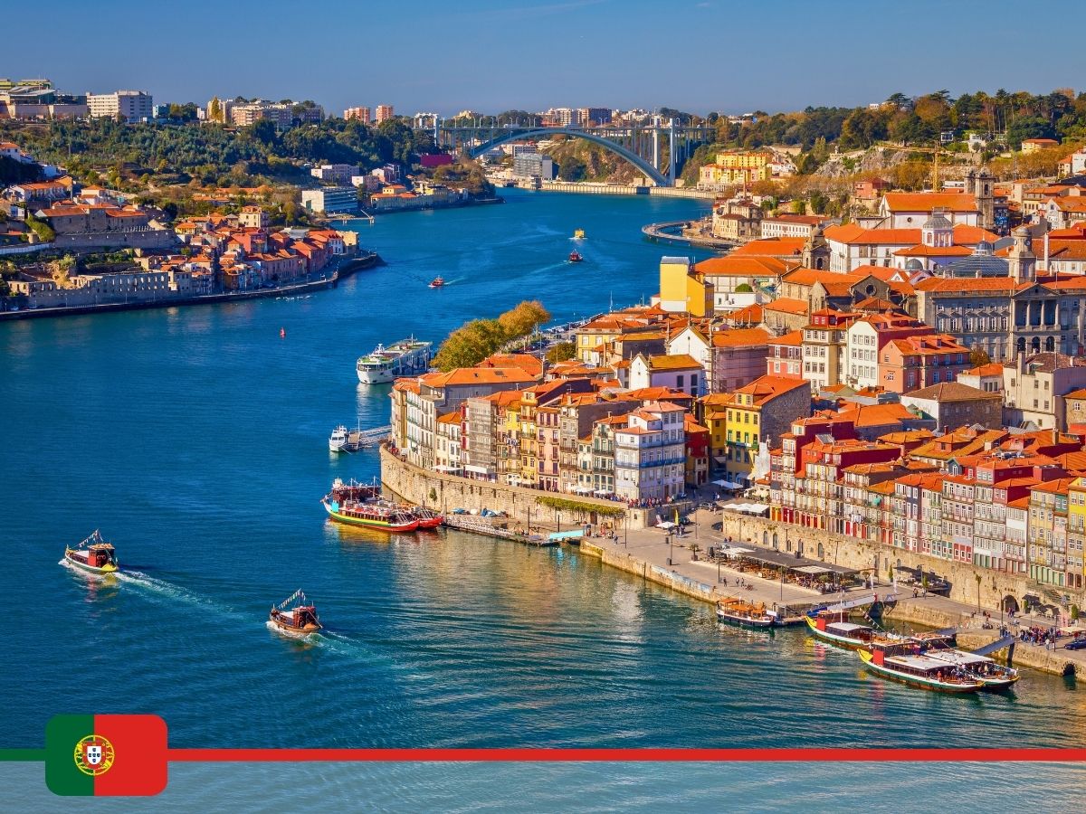 Aerial view of Porto, Portugal, showcasing a river with boats and colorful buildings along the waterfront, with the Portuguese flag at the bottom left corner.