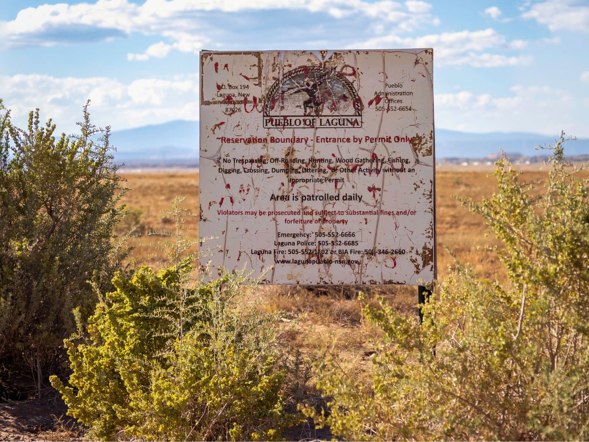 A weathered sign marks the boundary of the Pueblo of Laguna reservation, surrounded by desert plants and vast open landscape under a cloudy sky.